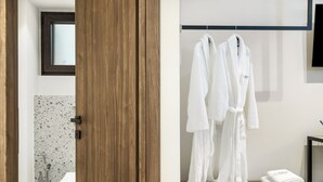 Une armoire en bois dotée d’une fenêtre en verre dépoli, une salle de bain avec un mur recouvert de carreaux à motifs, et une commode en bois sur laquelle sont suspendus et pliés deux peignoirs blancs.