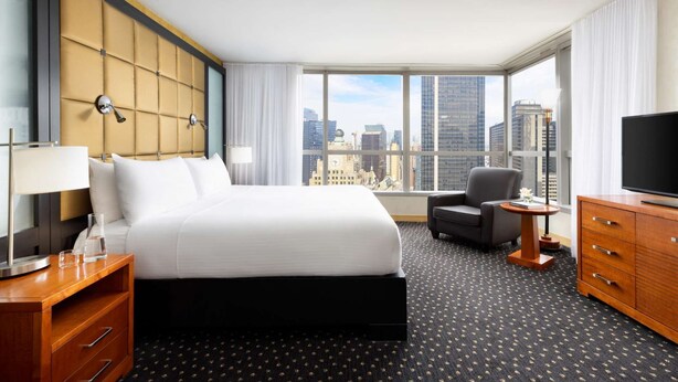 Habitación de hotel con una cama grande, un escritorio, una silla, un televisor y vistas a la ciudad.