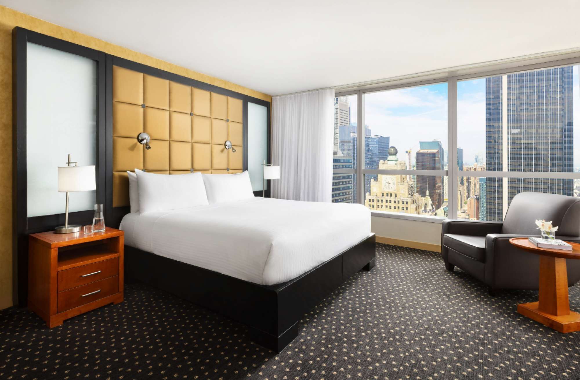 A hotel room with a large bed, a nightstand, a chair, and a view of the cityscape.