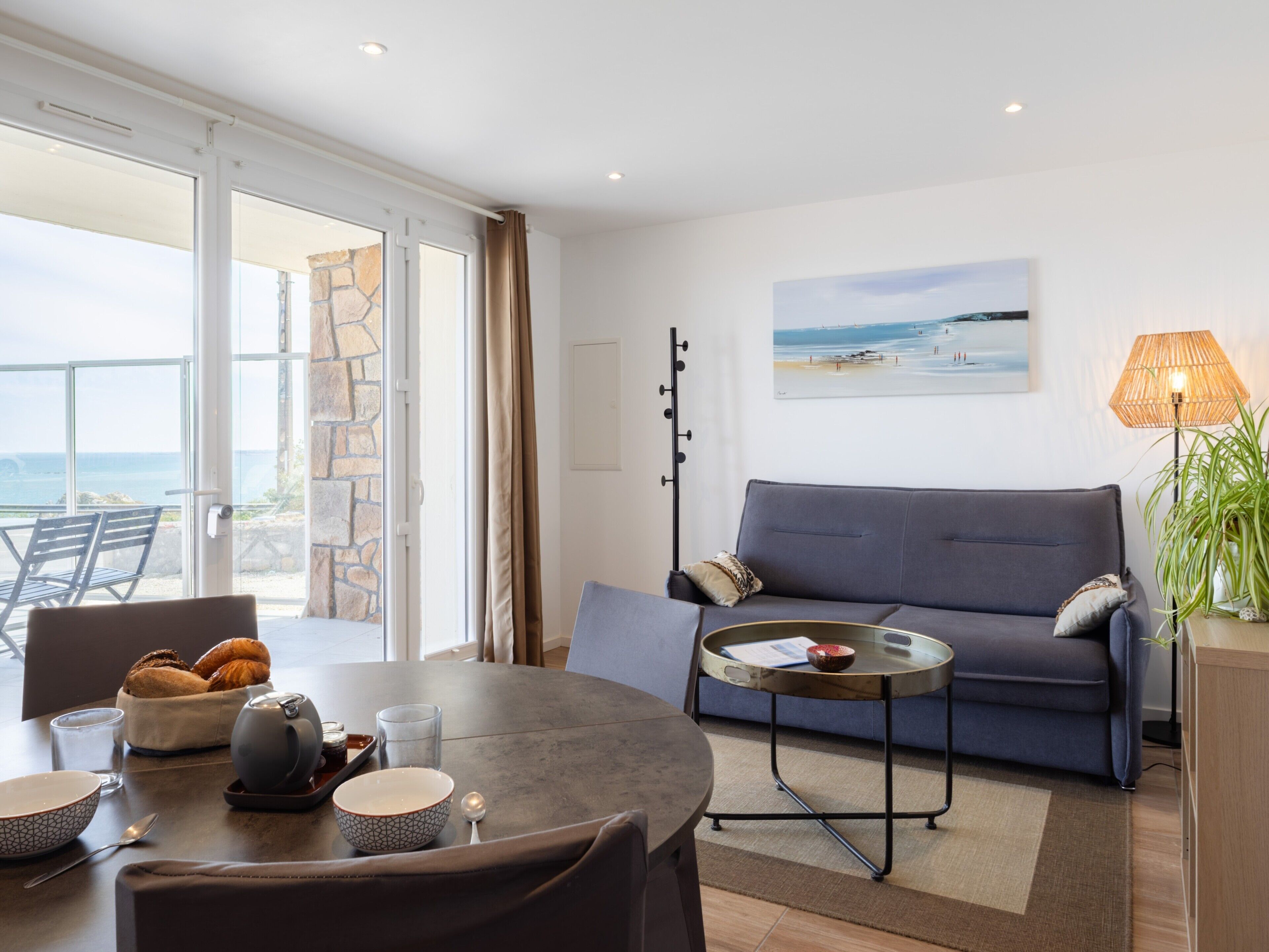 A modern living room with a sofa, a dining table with chairs, a coffee table, and a view of the ocean through large windows.