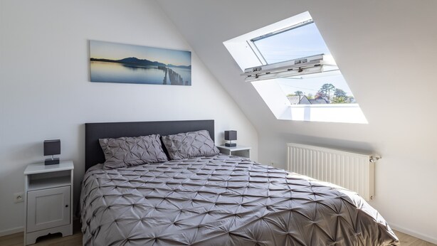 Une chambre à coucher comprenant un lit, une table de chevet, une commode, un radiateur et une lucarne.