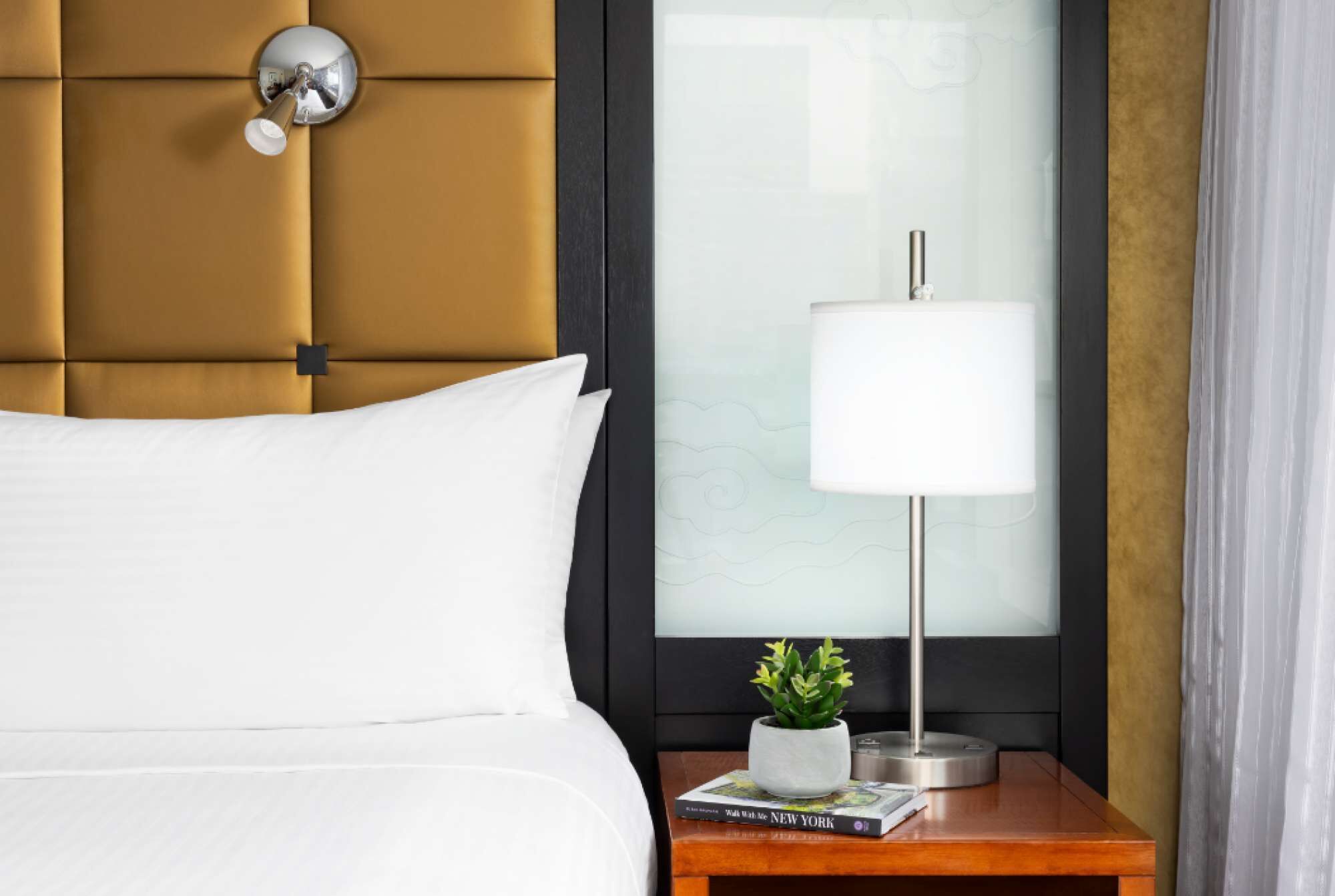 Habitación de hotel con cabecera capitonada, mesita de noche con lámpara, planta en maceta y un libro titulado "Walk With Me NEW YORK".