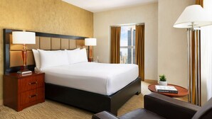 Ein Hotelzimmer mit einem großen Bett, zwei Nachttischlampen, einem Holznachttisch, einem kleinen Tisch mit einer Pflanze und Blick auf Gebäude durch das Fenster.