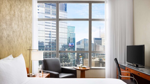 A hotel room with a large window, a bed, a desk, a chair, a TV, and a small table with a vase and books.