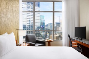 A hotel room with a large window, a bed, a desk, a chair, a TV, and a small table with a vase and books. - Electrifying City | Rockefeller l Ball Drop view (New York)