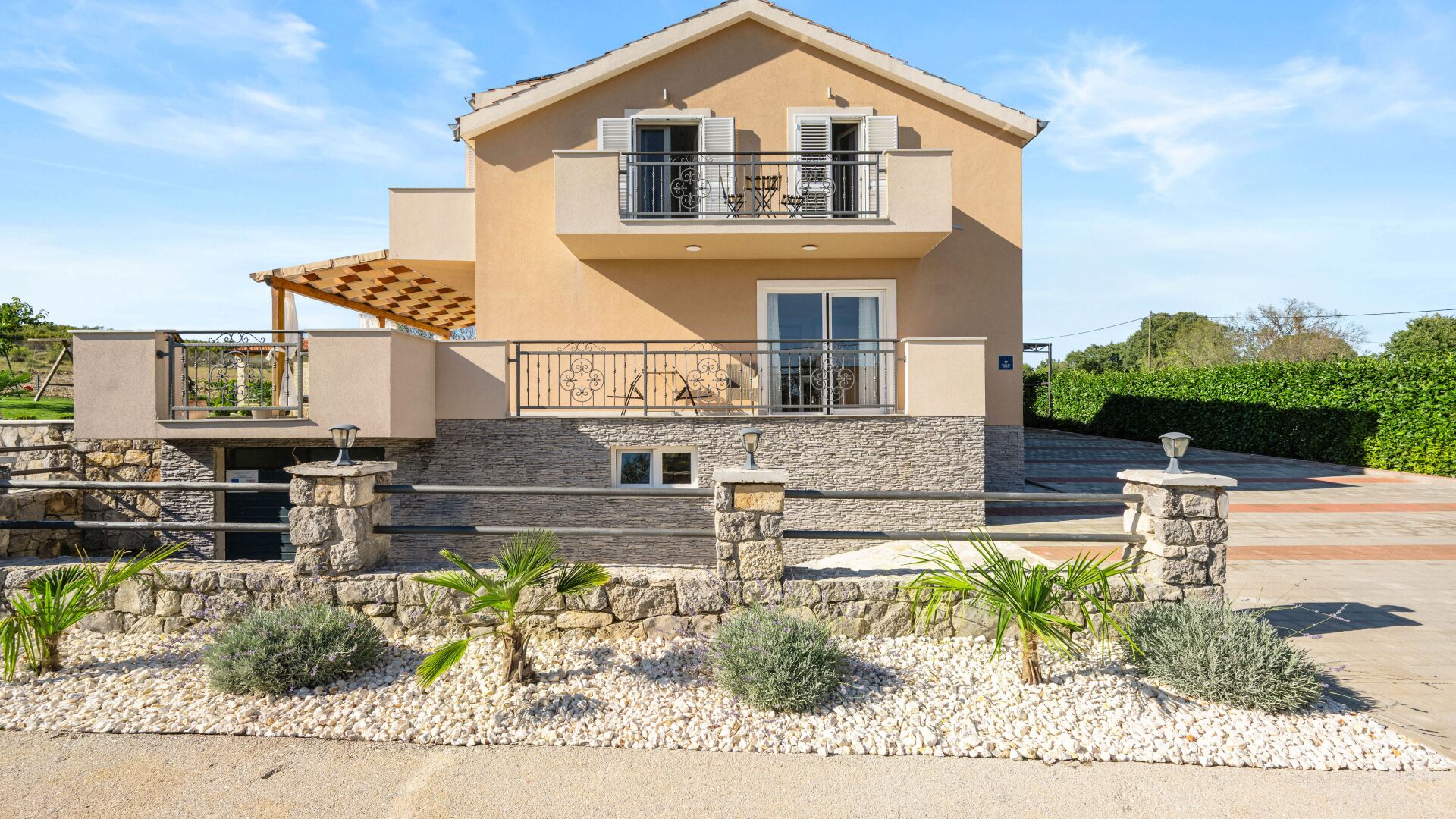 A two-story house with a stone facade, a balcony with metal railings, and a small garden with pebbles and plants.