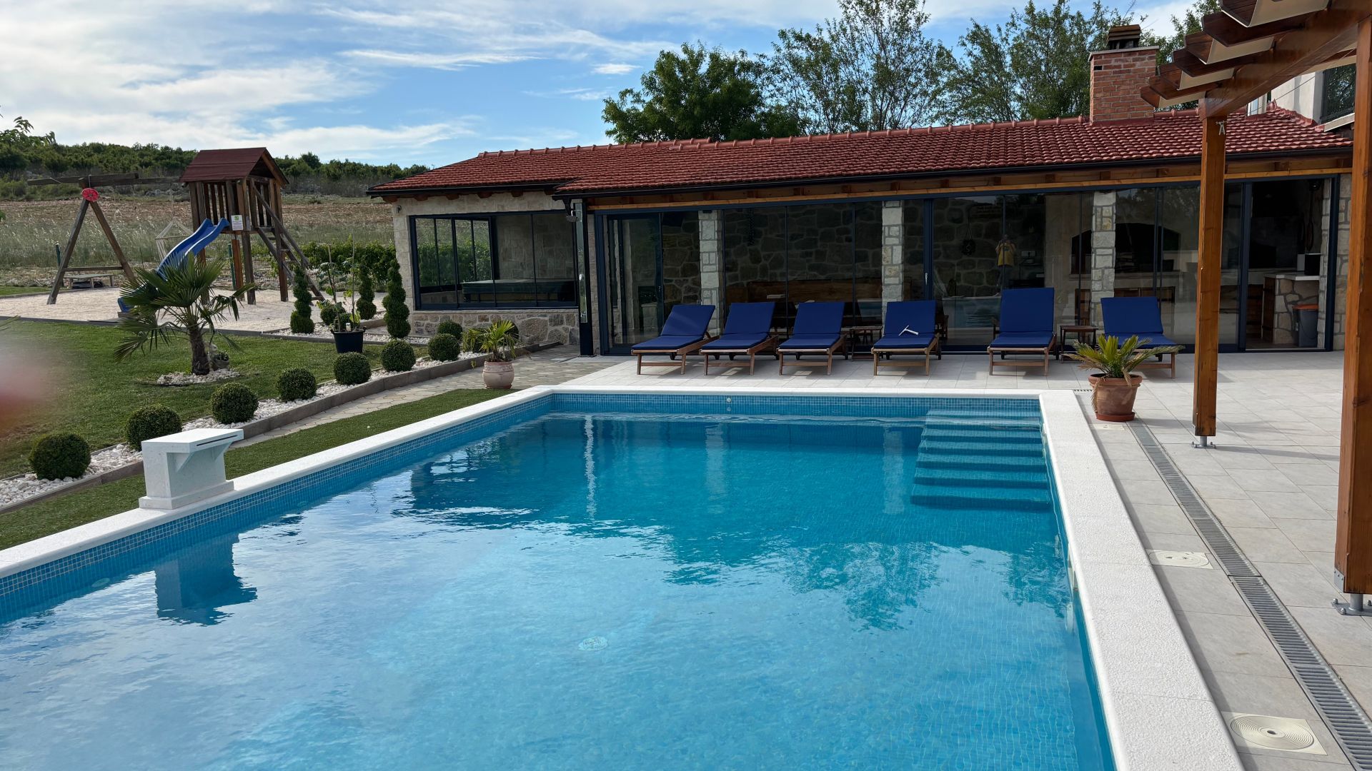 Piscina com espreguiçadeiras, área de recreação infantil e edifício de pedra com telhado de telhas vermelhas.