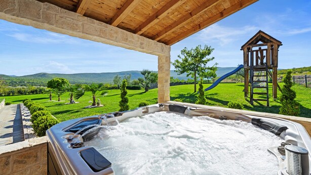 Ein Whirlpool mit Steinwand und Holzboden, der auf eine grüne Landschaft mit Spielplatz und Bergen in der Ferne blickt.