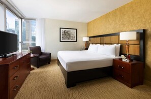 A hotel room with a large bed, a desk with a TV, a chair, a nightstand with a lamp, and a framed picture on the wall. - Sleepless City | Times Square. Fitness Center (New York)