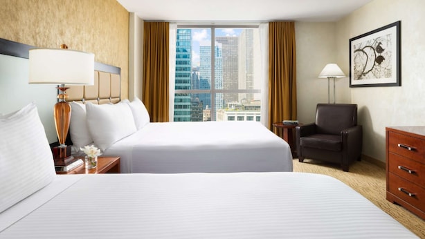 A hotel room with two beds, a chair, a desk, and a view of the cityscape. - Double Deluxe | Times Square l City View (New York)