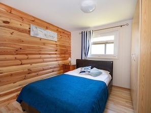 A bedroom with a wooden log wall, a bed with a blue bedspread, a nightstand with a lamp, and a window with curtains. - Comfortable Holiday Homes, Jacuzzi, Summer Pool (Postomino)