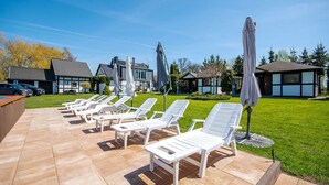 A poolside area with lounge chairs and umbrellas, a grassy lawn, and buildings in the background. - Comfortable Holiday Homes, Jacuzzi, Summer Pool (Postomino)
