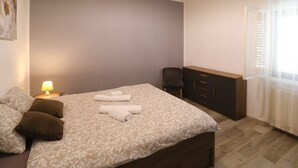 Ein Schlafzimmer mit einem Bett, Kissen, einem Nachttisch mit Lampe, einem Sessel, einer Kommode und einem Fenster mit Jalousien.