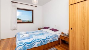 A bedroom with a bed, a nightstand, a wooden wardrobe, a window with a view, and a hanging light fixture. - Apartman FLORIS with AC and WIFI (Punat)
