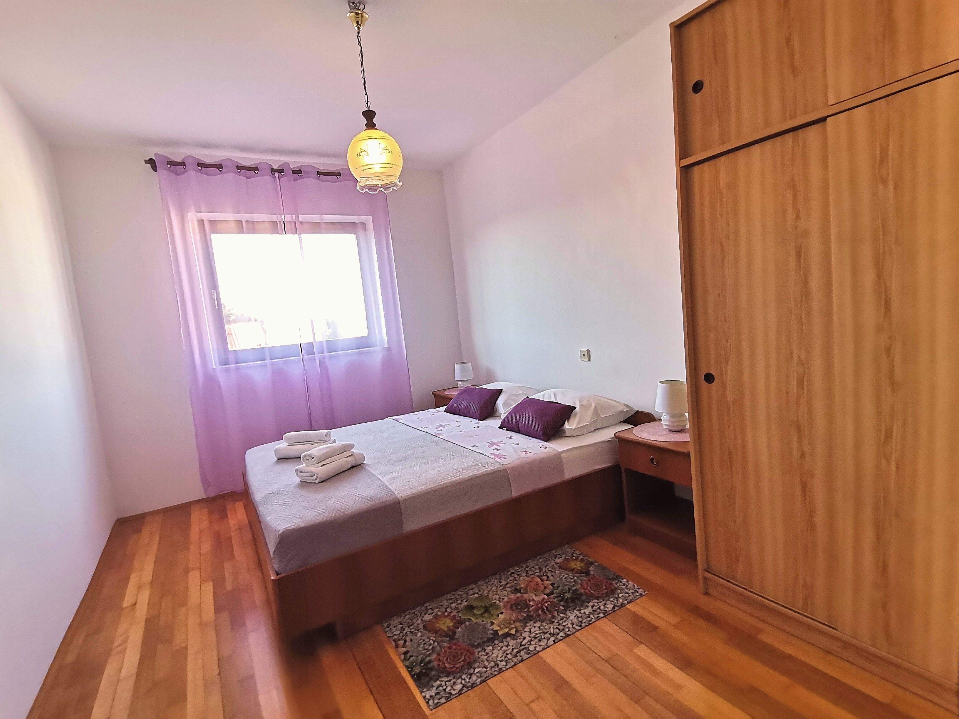 A bedroom with a bed, bedside lamps, a wooden wardrobe, and a window with curtains.