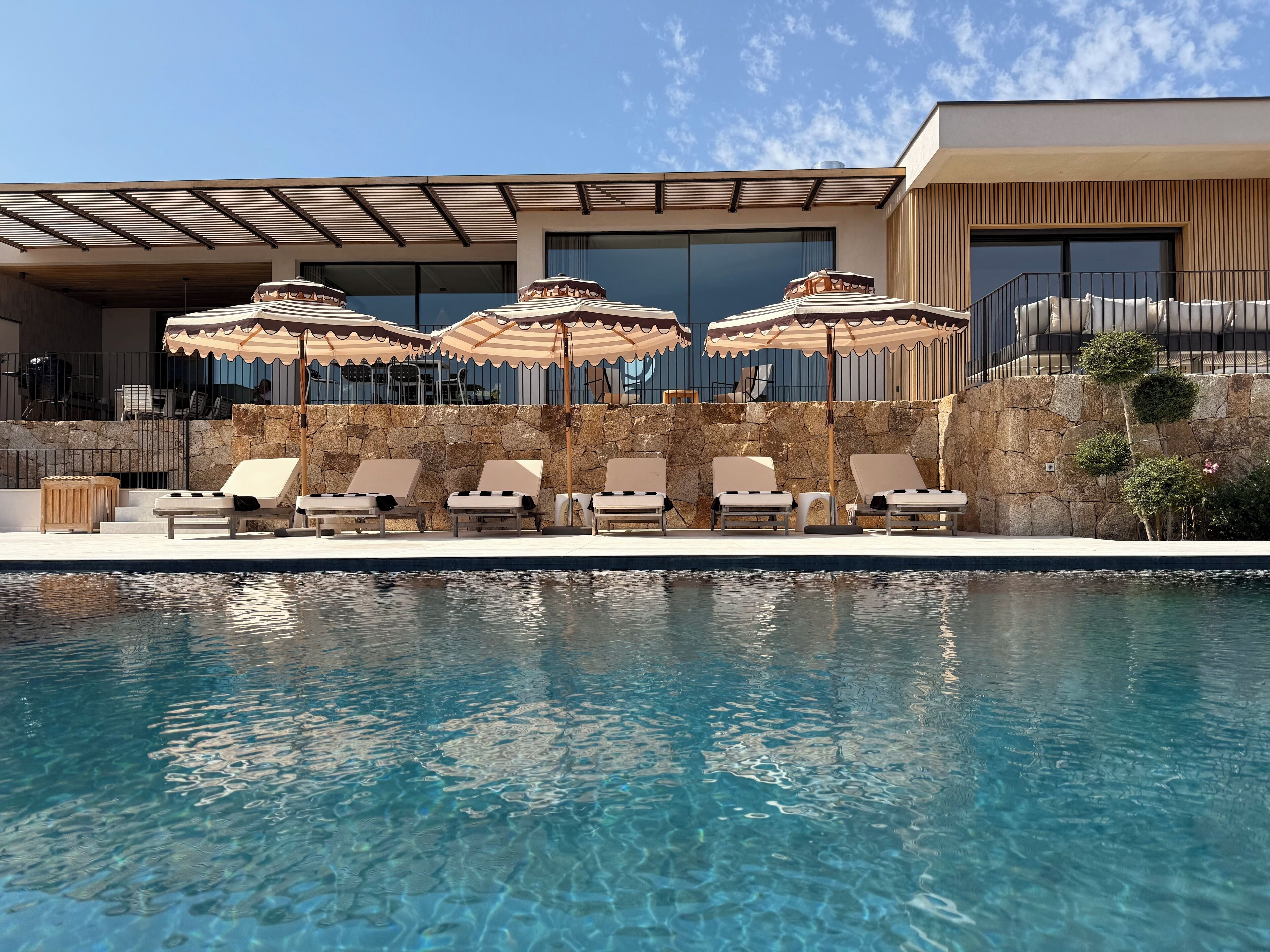 A poolside area with lounge chairs and umbrellas, a stone wall, and a modern building with large windows.