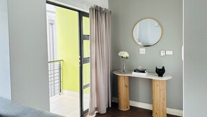 Ein Zimmer mit einer Schiebetür zu einem Balkon, einem runden Spiegel an der Wand, einem kleinen Holztisch mit einer Vase und Büchern sowie einem grauen Sofa.