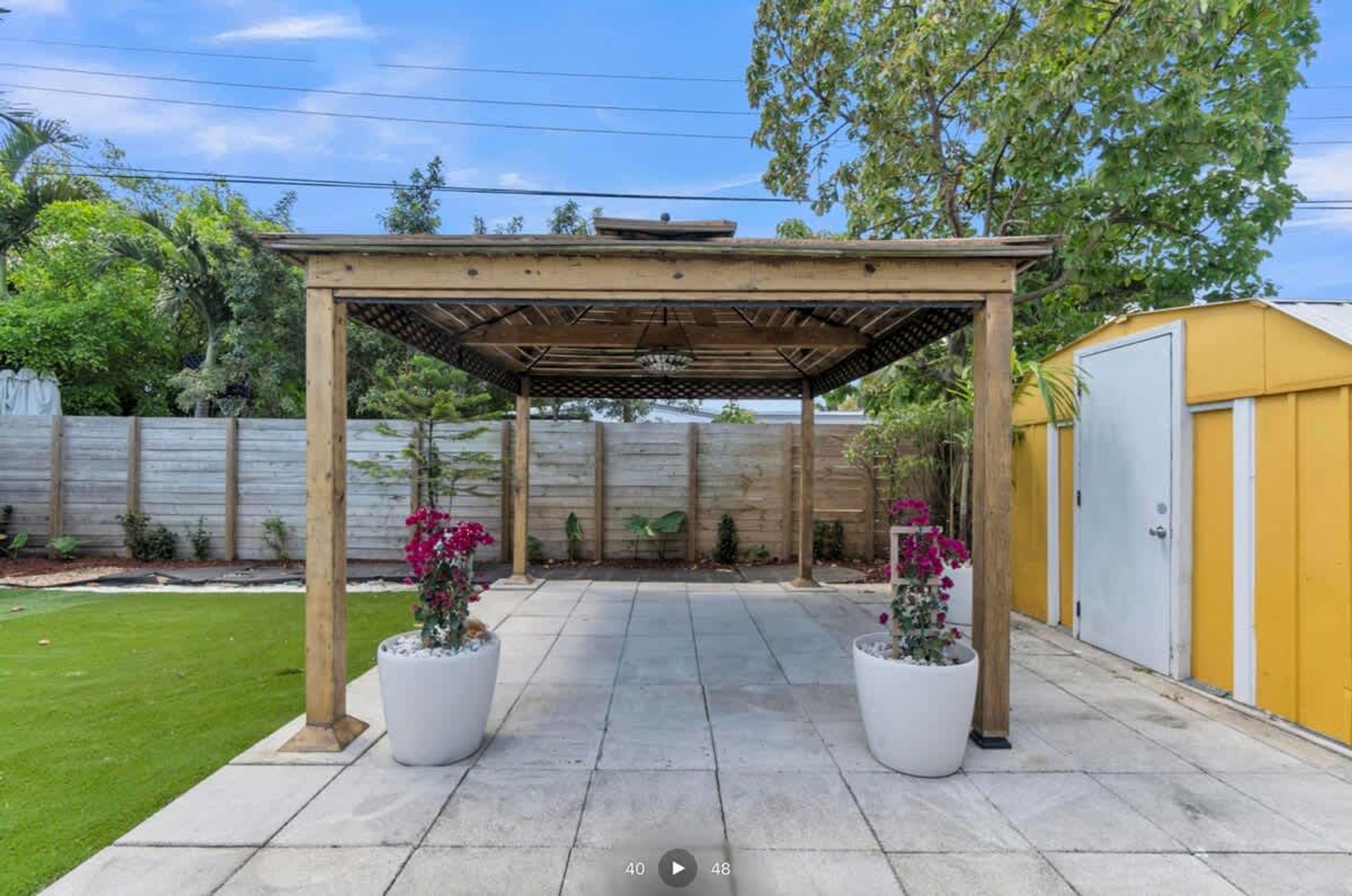 Un patio abrité doté d’une pergola, de plantes en pot et, au fond, d’un bâtiment jaune.