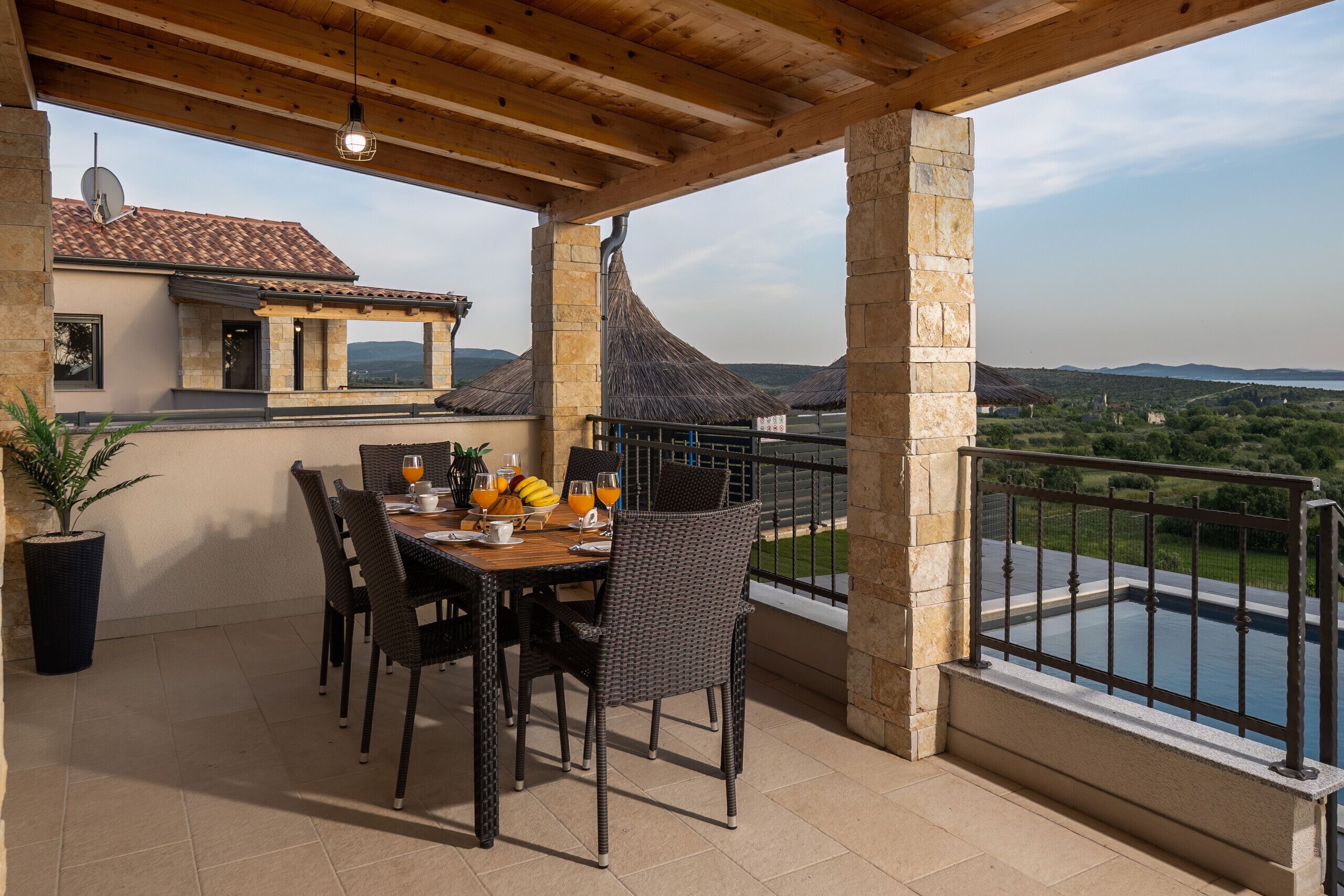 Un espace extérieur abrité, aménagé pour prendre les repas, avec une table pour deux, une plante en pot et une vue sur une piscine et un jardin.