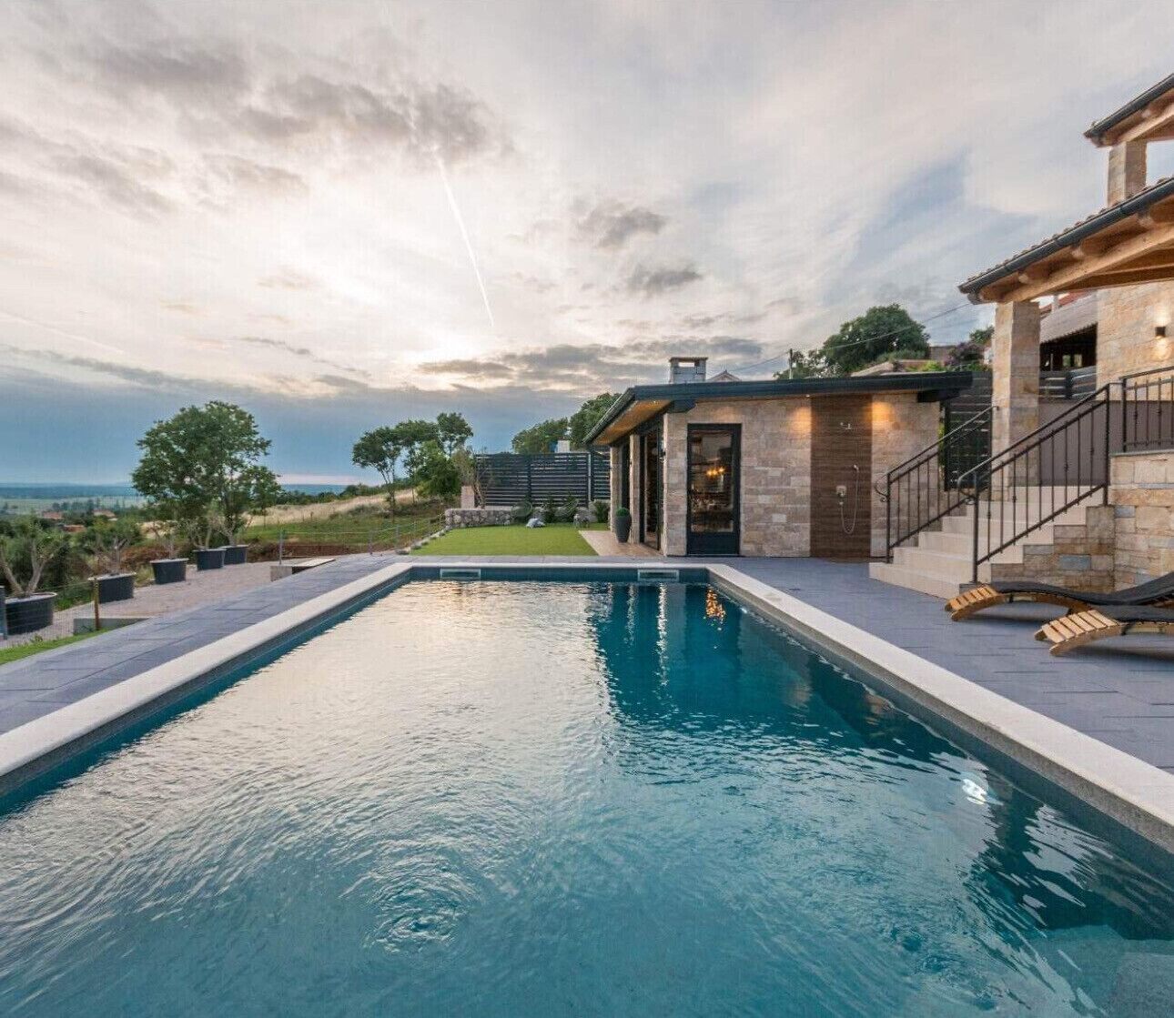Une piscine avec une terrasse en pierre, une maison moderne dotée d’une façade en pierre et une vue sur le paysage.