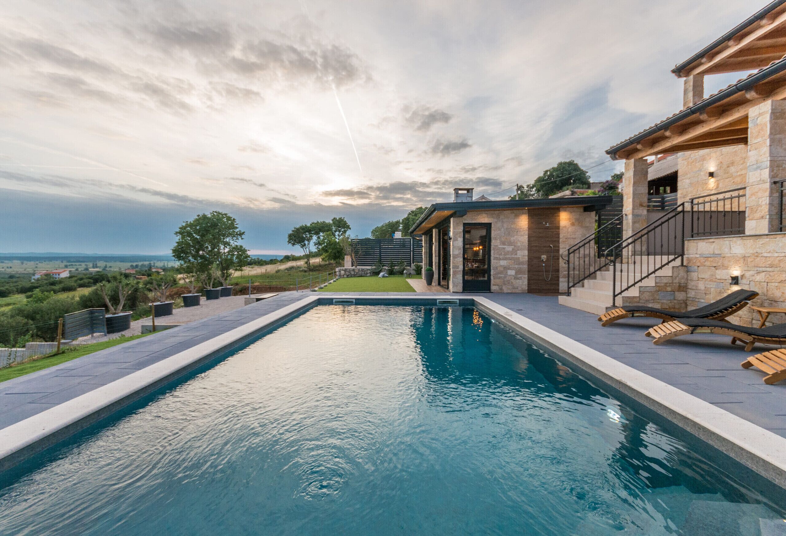 Une piscine rectangulaire entourée d’une terrasse en pierre, un espace en bois avec des chaises longues et un bâtiment en pierre doté d’un escalier.
