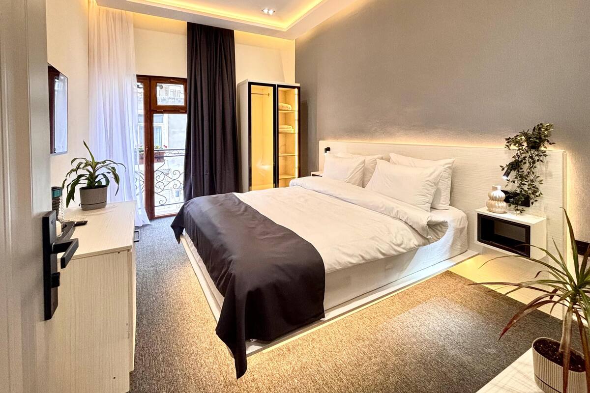 A modern bedroom with a large bed, a bedside table, a nightstand, a potted plant, and a view of the outdoors.