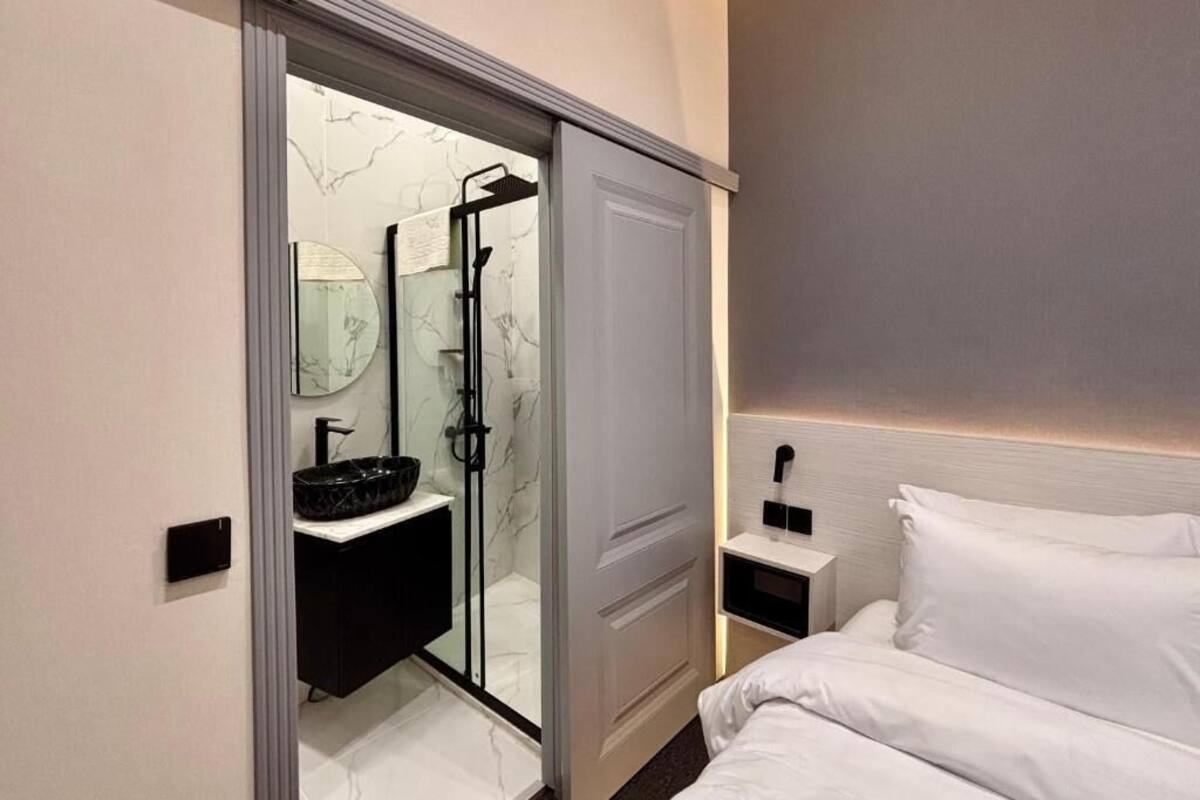 A modern bathroom with a glass shower enclosure, a white sink, and a bedside table with a lamp.