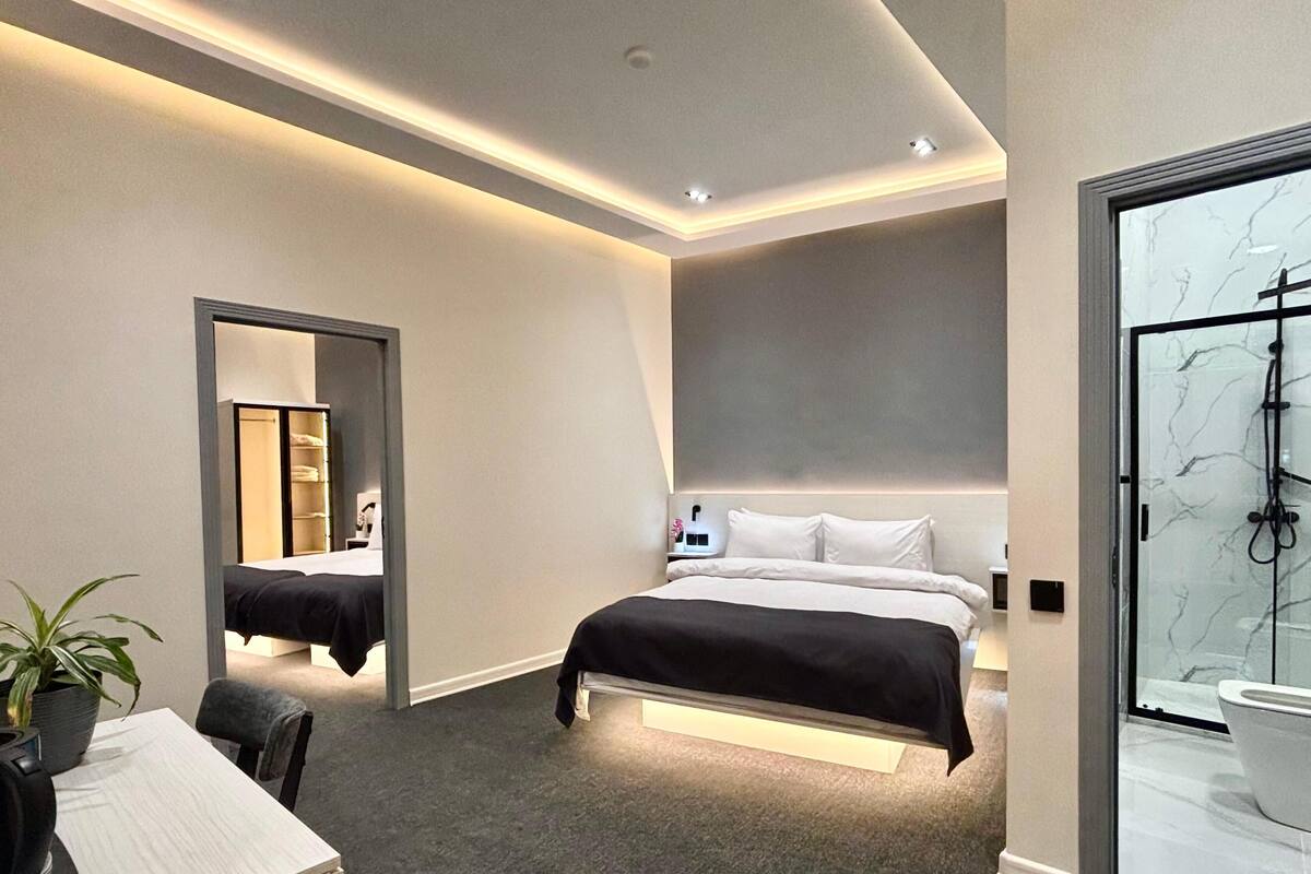 A modern hotel room with a large bed, a desk with a chair, a shower area, and a mirror.