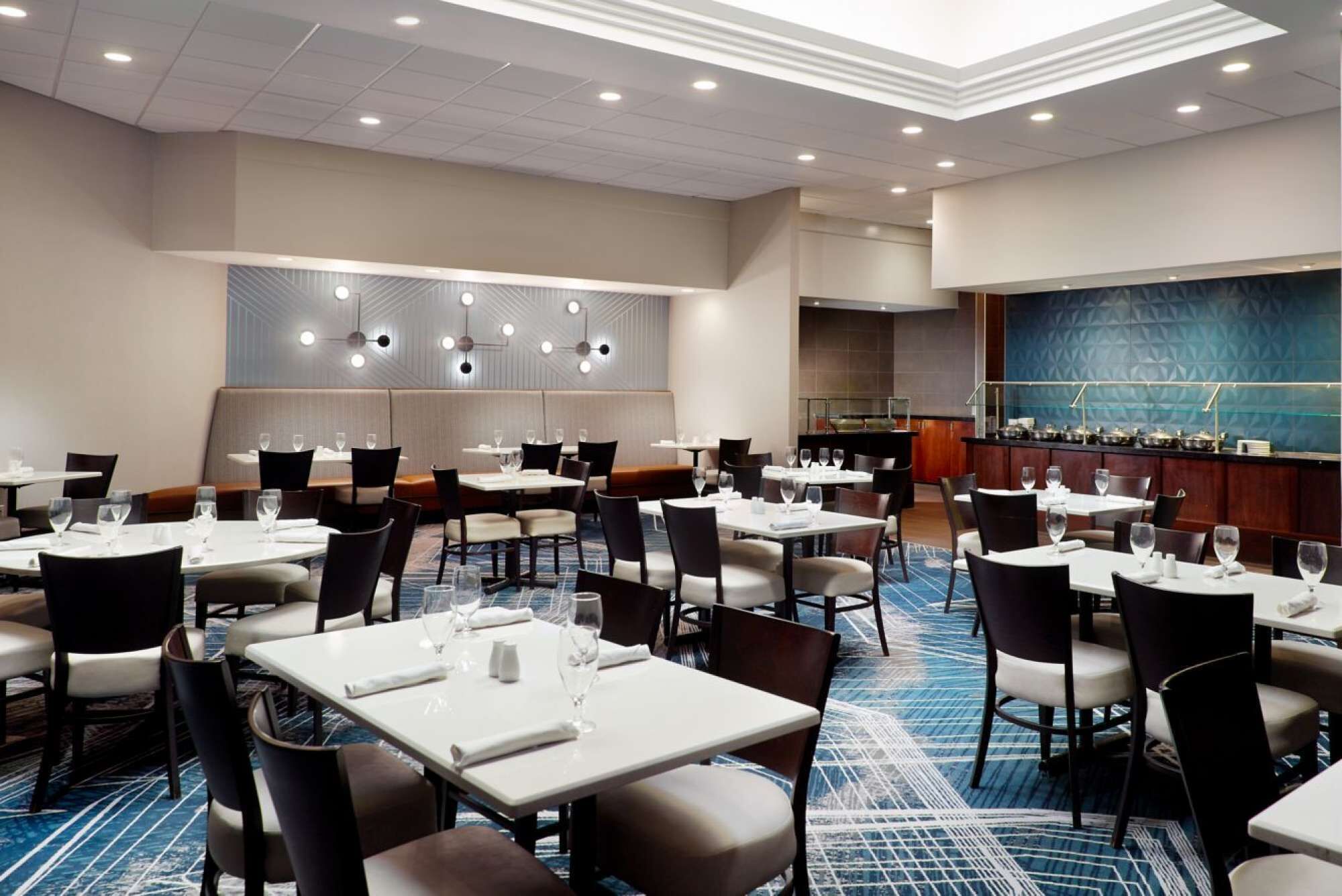 A hotel dining area with multiple tables and chairs, a bar area, and a patterned carpet.