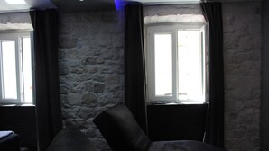 Ein Zimmer mit Steinwänden, zwei mit Vorhängen versehenen Fenstern und einem dunkelbraunen Sofa.