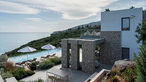 A modern building with a stone facade, a pool area with lounge chairs, and a view of the sea.
