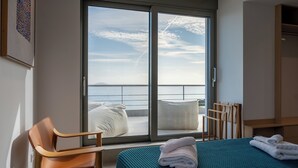 A hotel room with a bed, a chair, a view of the sea through a sliding door, and a wooden wardrobe.