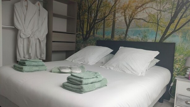 Uma cama bem arrumada com roupas de cama brancas e toalhas verdes, uma cabeceira com um mural de tema natural e um roupão pendurado em um suporte.