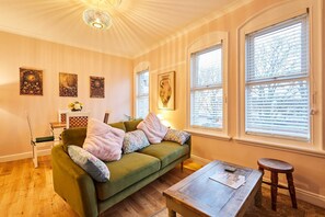 Un salon chaleureux avec un canapé vert, une table basse en bois et un petit tabouret. Il y a trois fenêtres équipées de stores, un lustre et des œuvres d’art accrochées au mur.