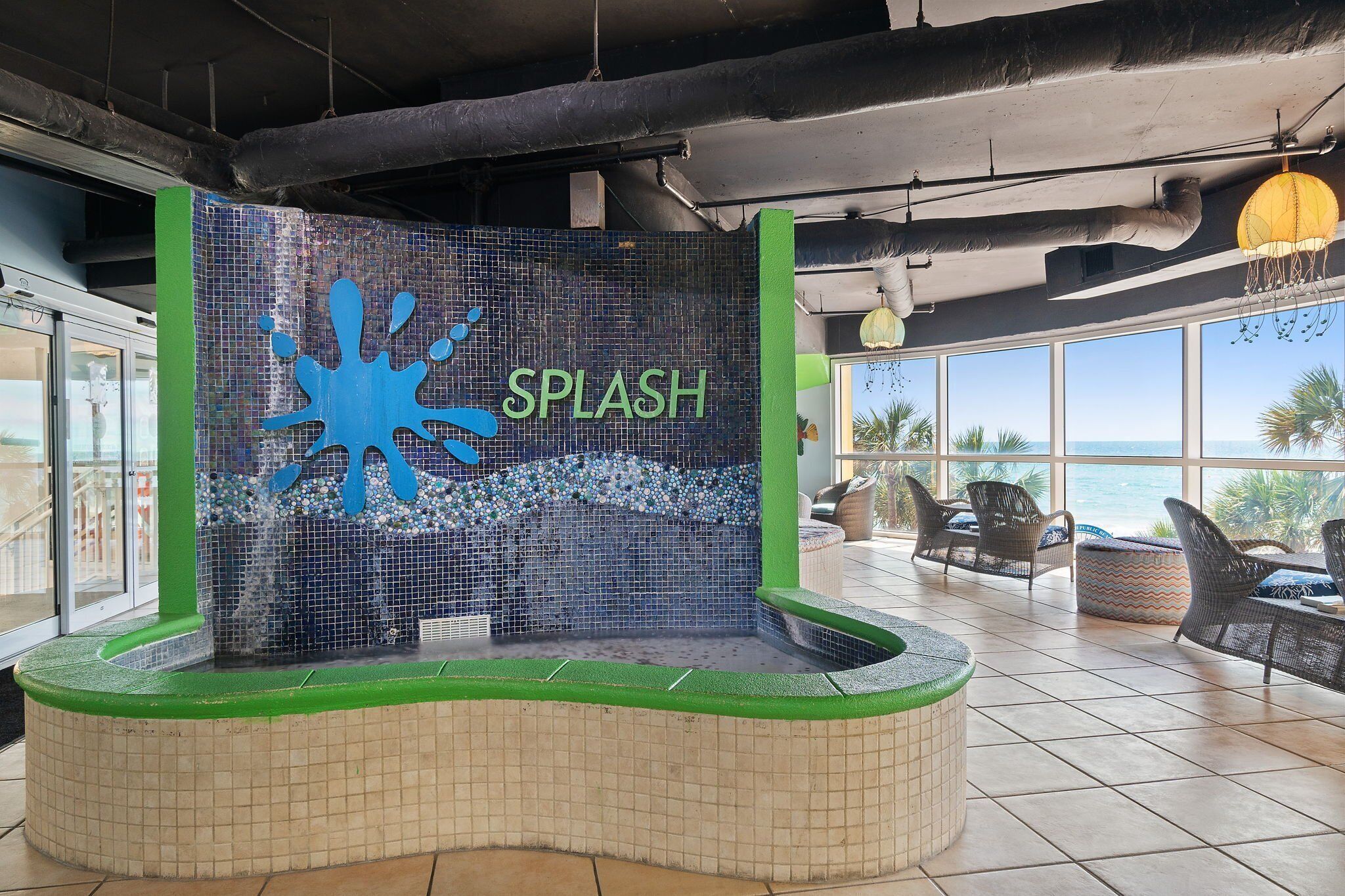 Una fuente con agua y la palabra "SPLASH" en el centro, rodeada de un área con azulejos, sillas y vistas al océano.
