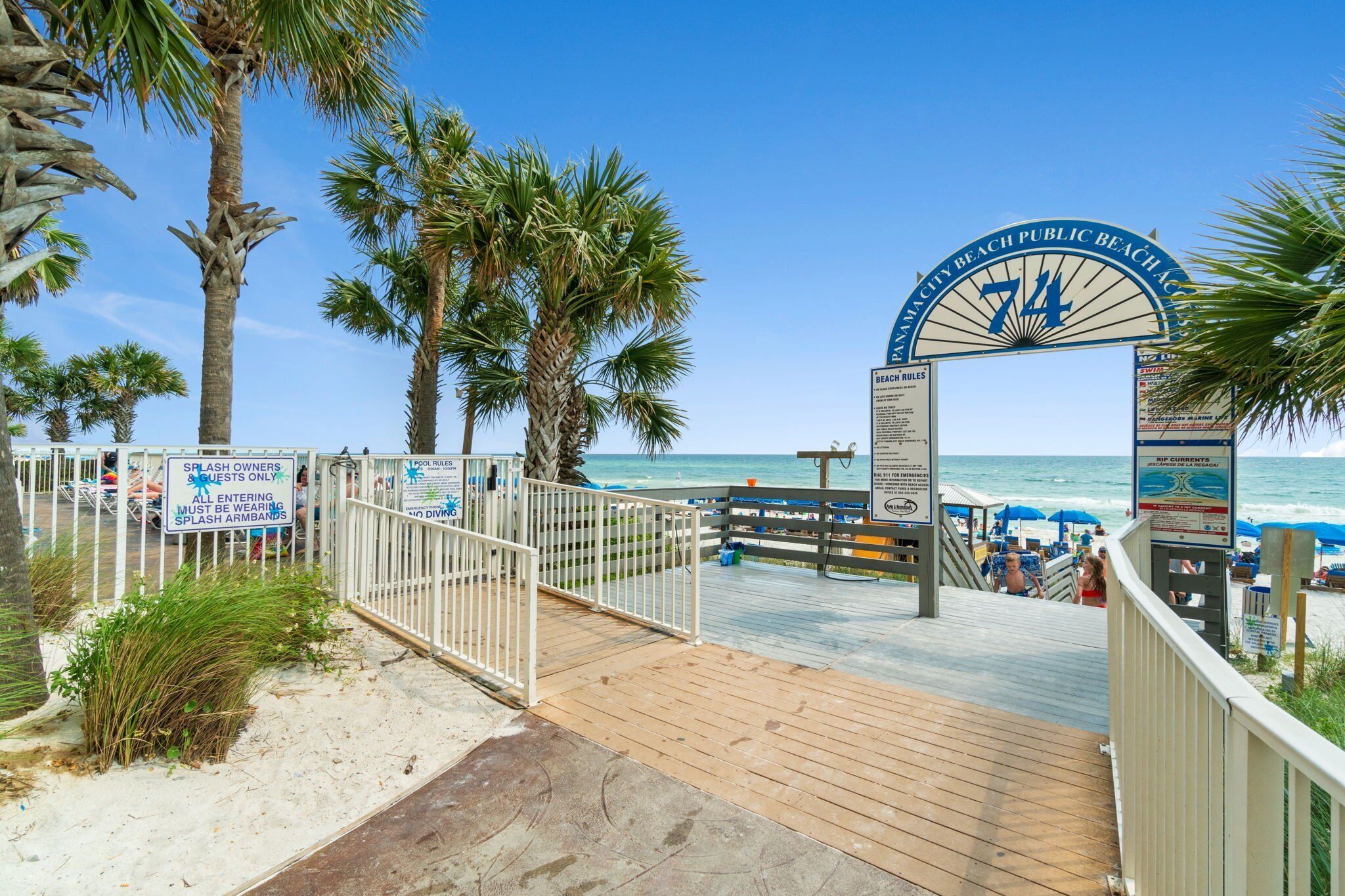 A wooden boardwalk leading to a beach with palm trees and a sign indicating '72' for the beach number.