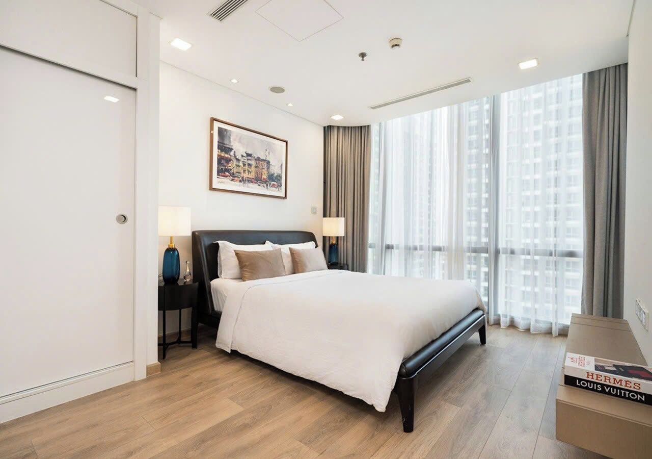 A modern hotel room with a large bed, a nightstand with lamps, a framed picture on the wall, and a view of buildings through the window.