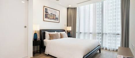 A modern hotel room with a large bed, a nightstand with lamps, a framed picture on the wall, and a view of buildings through the window.