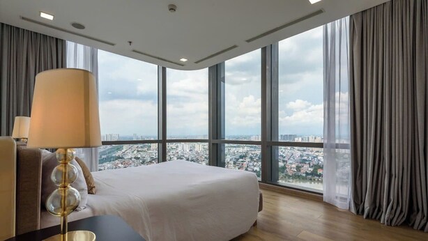 Ein Hotelzimmer mit einem großen Bett, einem Nachttisch mit Lampe und Blick auf eine Stadtkulisse durch große Fenster.