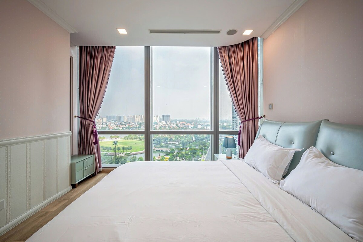 Ein Schlafzimmer mit einem großen Bett, einem Nachttisch und Blick auf eine Stadtlandschaft durch raumhohe Fenster.