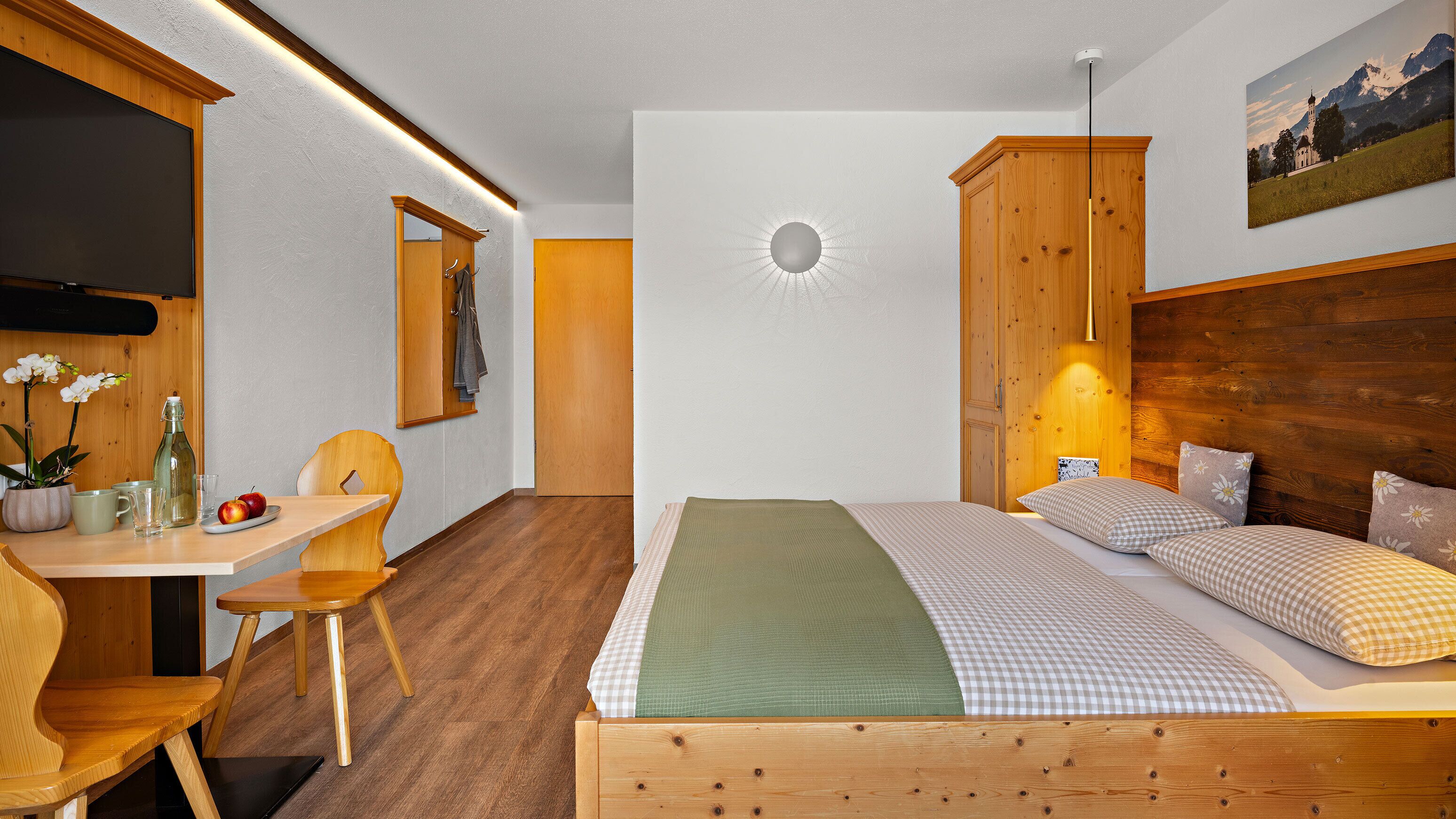 A bedroom with a wooden bed, a desk with a chair, a small table with a potted plant, and a painting on the wall.