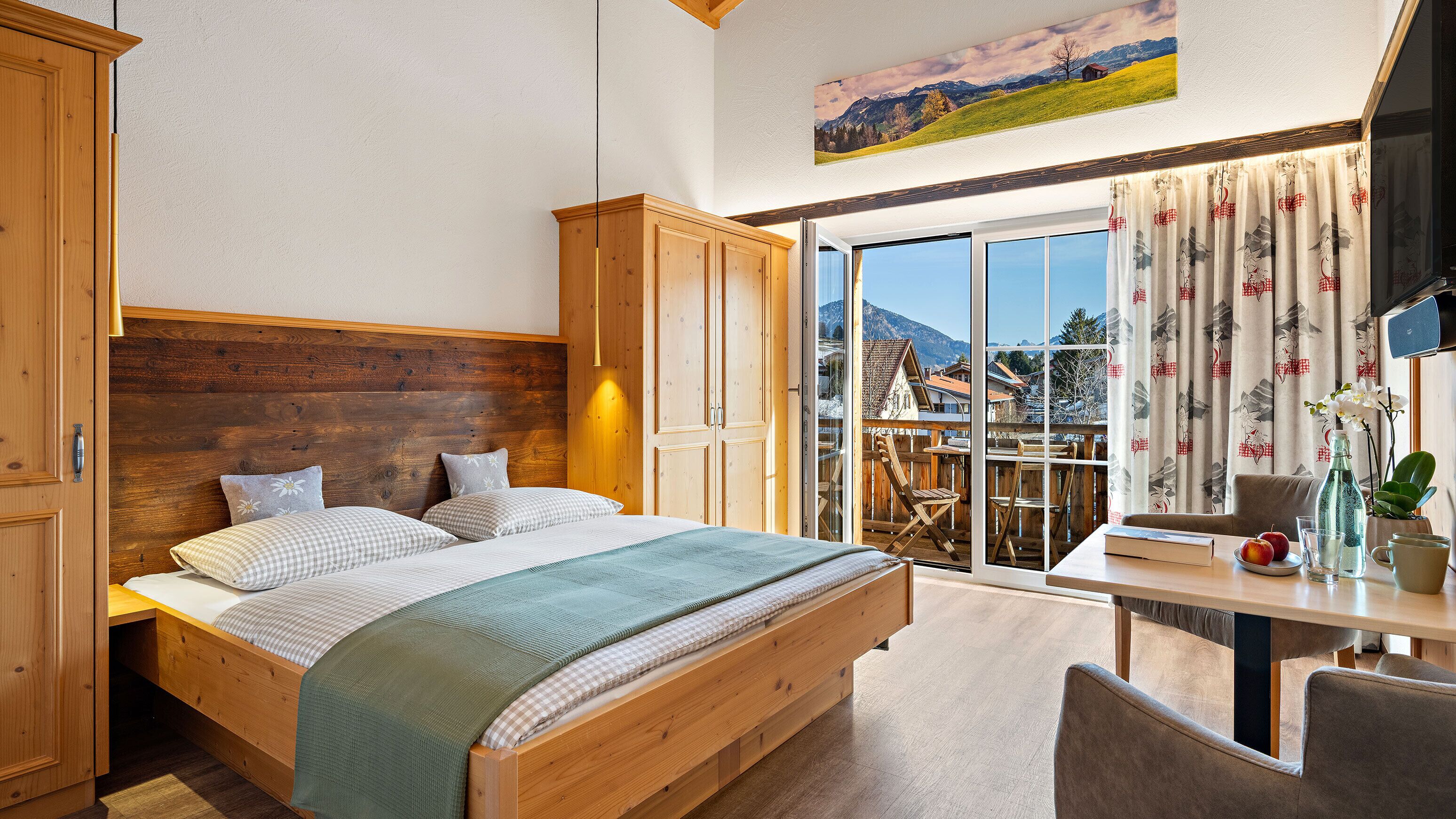 Une chambre avec un lit en bois, une armoire en bois, un balcon avec une table et des chaises, et un tableau représentant un paysage accroché au mur.