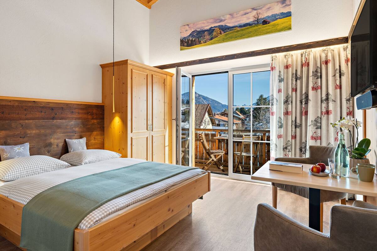 A bedroom with a wooden bed, a wooden wardrobe, a balcony with a table and chairs, and a painting of a landscape on the wall.