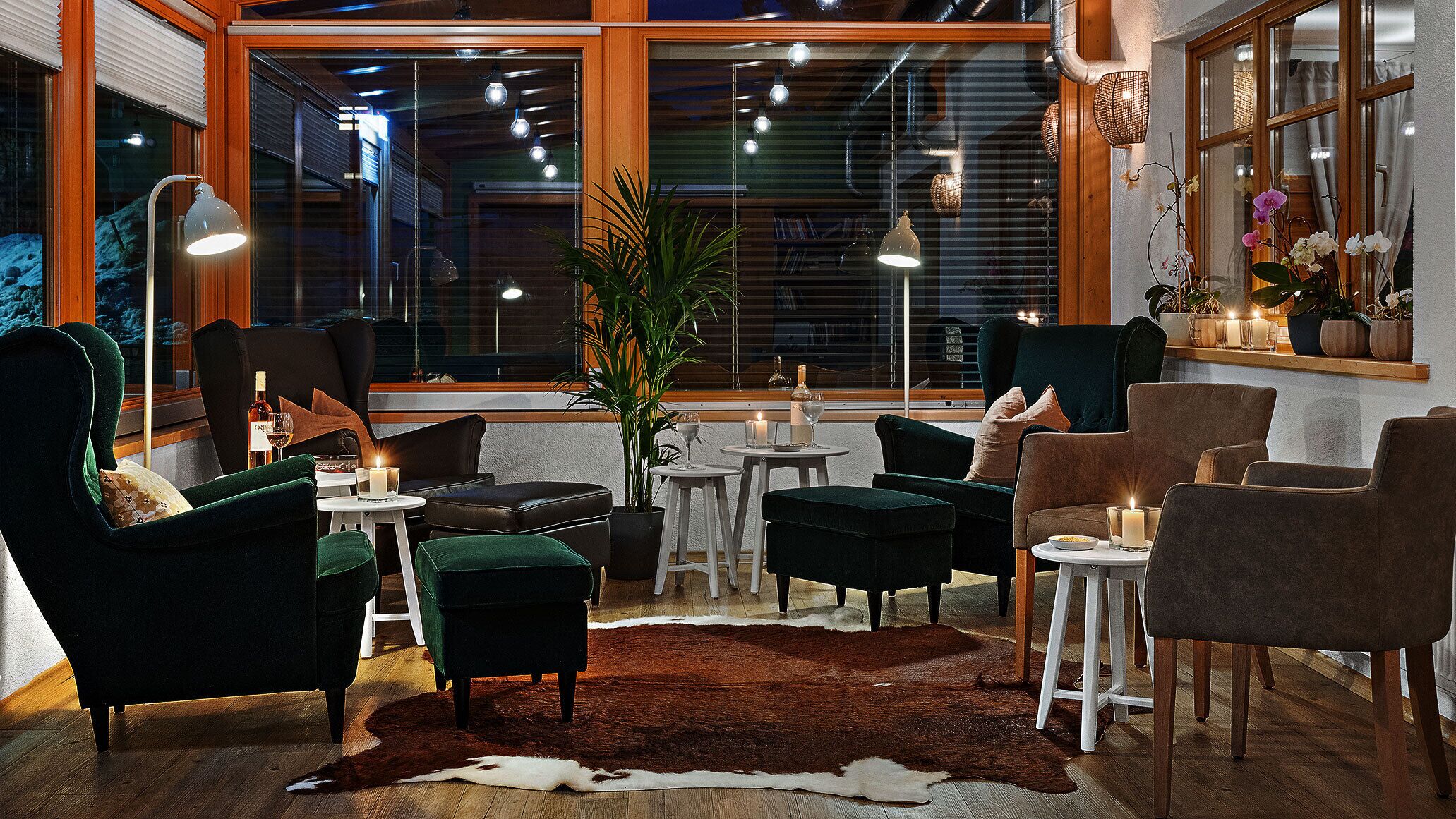A cozy lounge area with dark green armchairs, a leather sofa, and a cowhide rug. There are small tables with candles and drinks, and a large window with blinds and hanging lights.