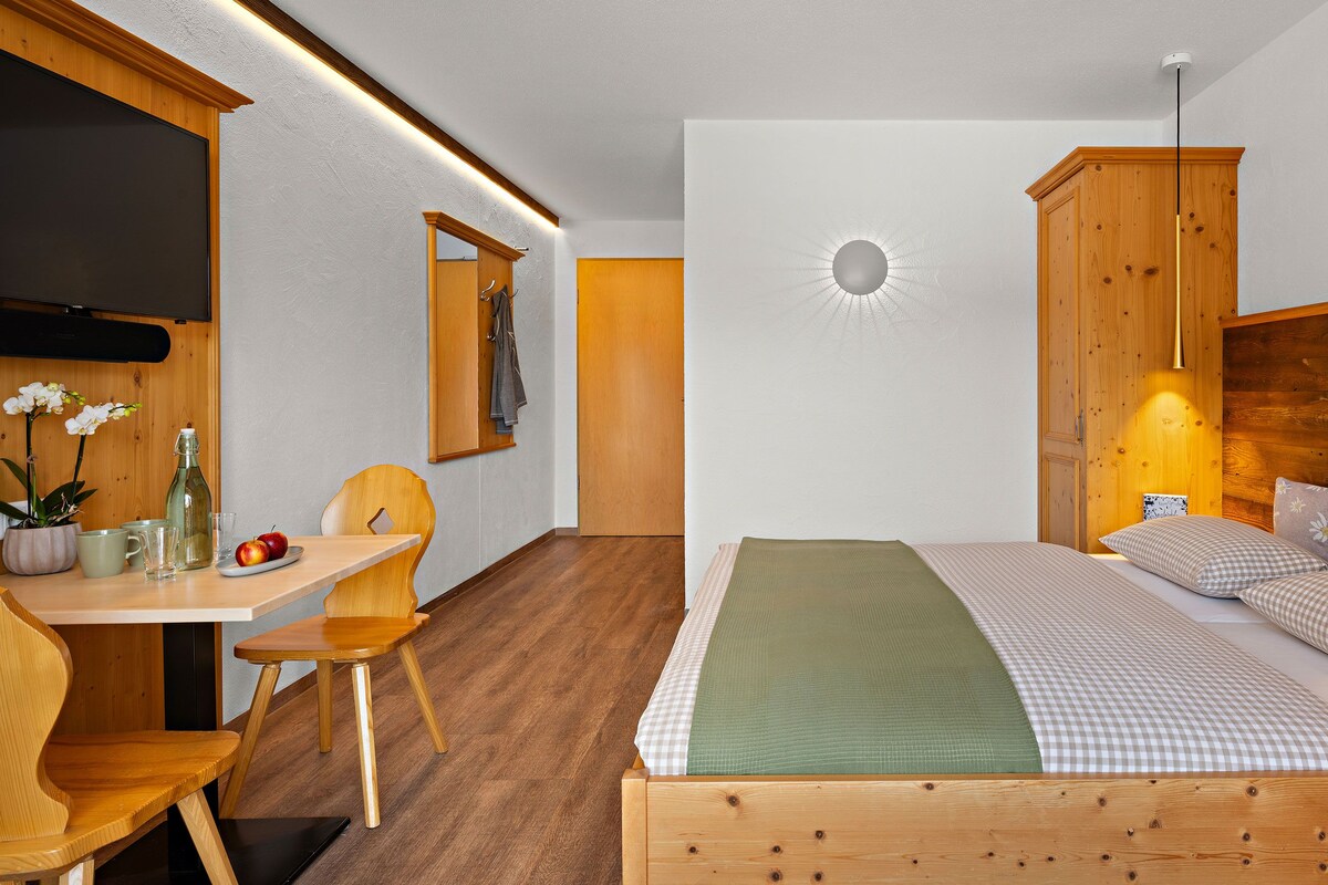 Une chambre avec un lit en bois, un bureau avec une chaise, une petite table sur laquelle est posée une plante en pot, et un tableau accroché au mur.