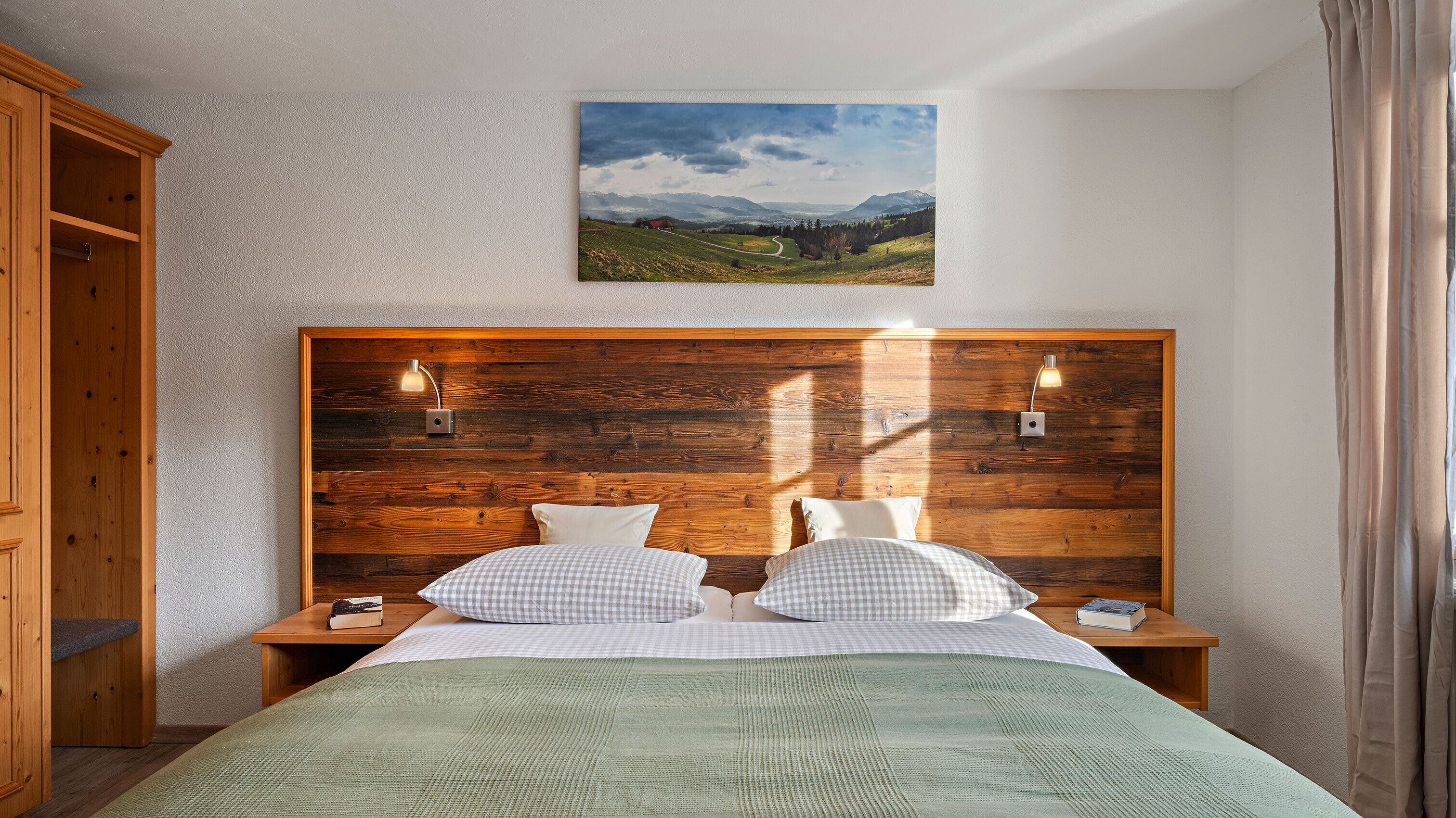 A bedroom with a wooden headboard, two bedside tables, a bed with checkered bedding, a green blanket, and a landscape painting above the headboard.