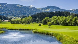 Un campo de golf con un estanque, árboles y montañas al fondo.