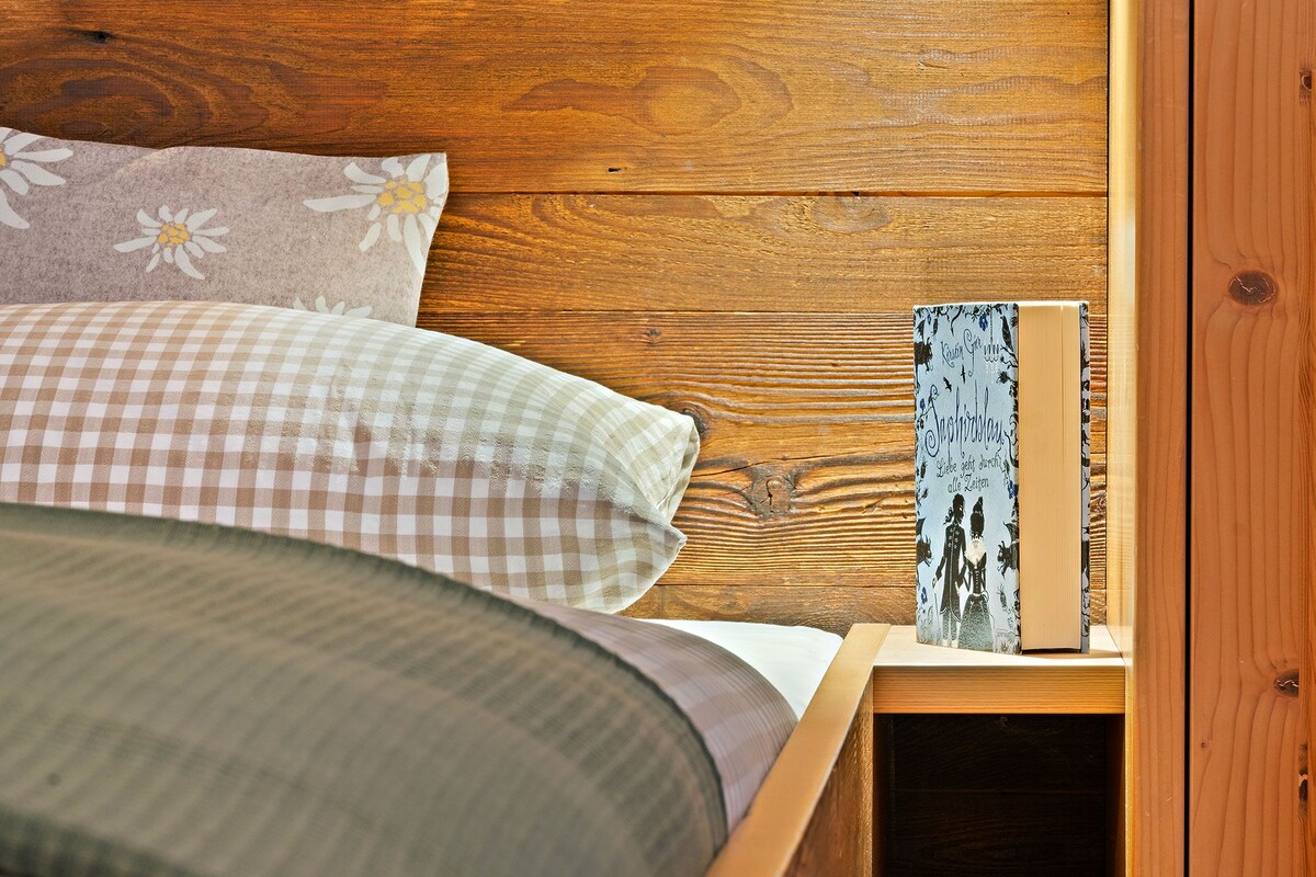 A wooden bed with a checkered blanket, pillows with floral patterns, and a bedside table with a book.