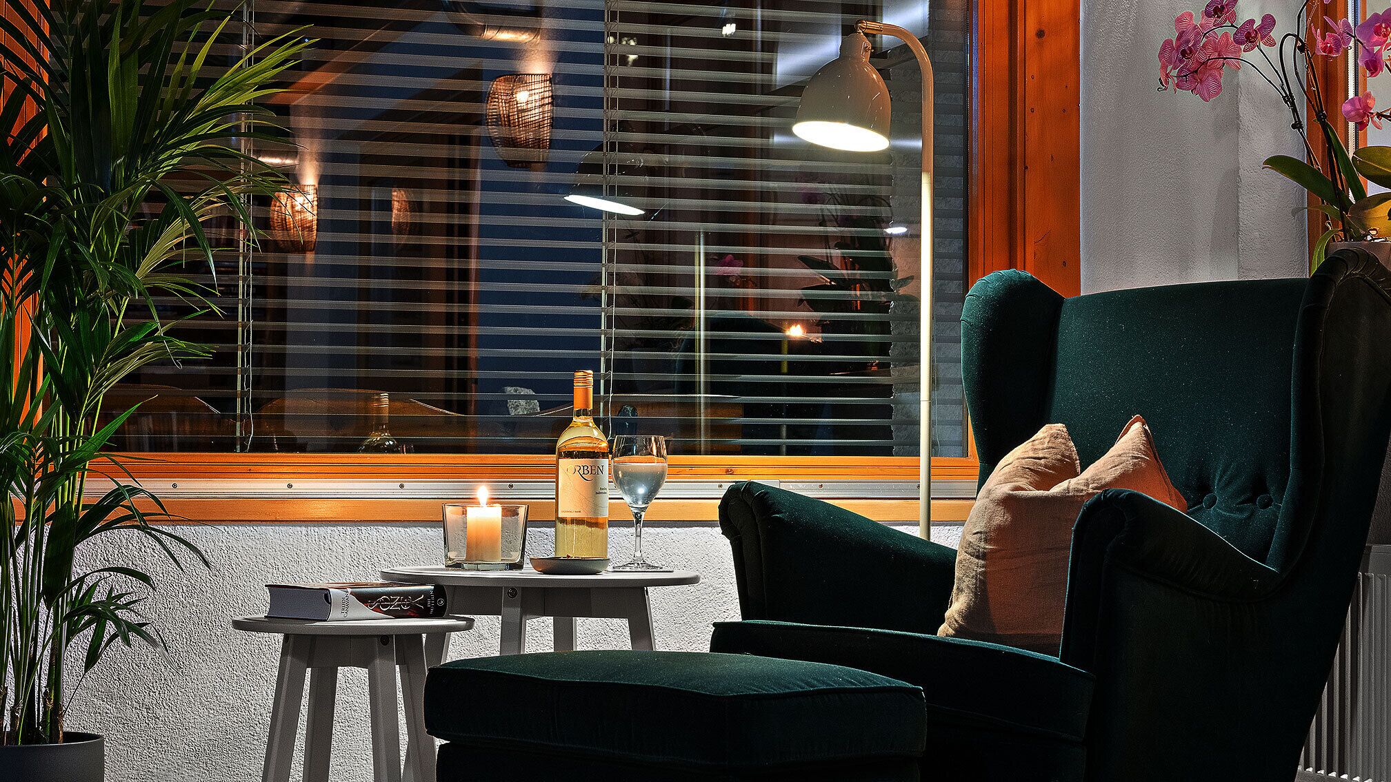 Un rincón acogedor con un sillón verde oscuro, una mesita con una vela, una botella de vino y una copa.