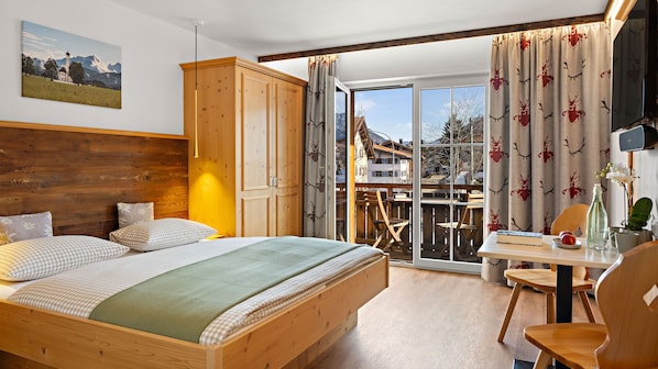 A bedroom with a wooden bed, a view of a balcony, and a dining area with a table and chairs. - Hotel Bergsonne Allgäu (Sonthofen)